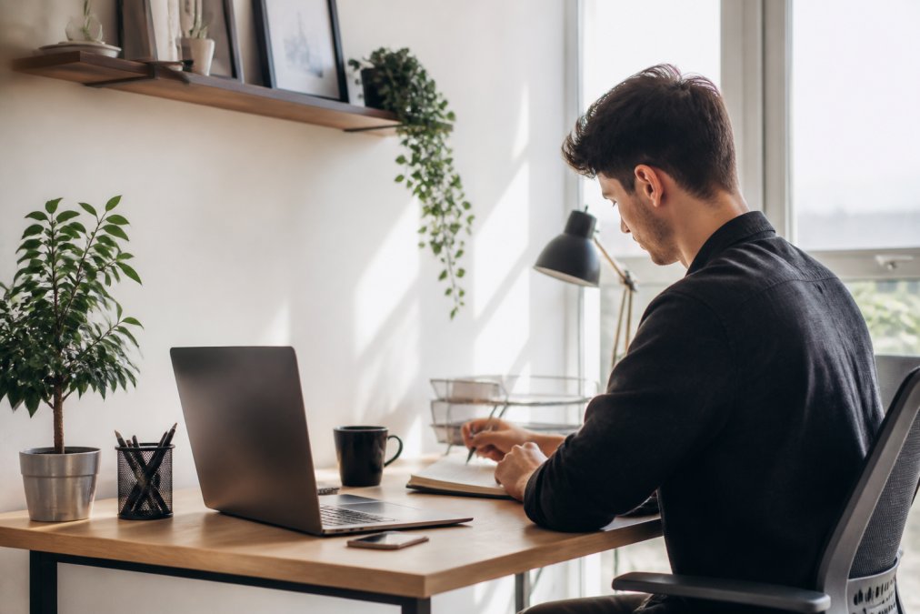 Junger Freelancer sitzt an einem modernen Schreibtisch und gestaltet eine Website mit einem visuellen Seiteneditor auf dem Monitor, helles, professionelles Büro-Umfeld mit natürlichem Licht von rechts