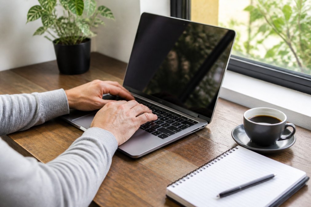 Close-up van handen die op een laptop typen met het WordPress dashboard zichtbaar op het scherm, koffiekopje en notitieblok op het bureau ernaast, zachte daglichtverlichting