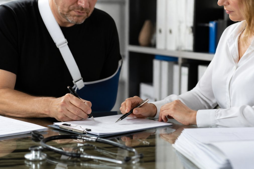 Close-up of hands completing workers compensation claim forms on a desk, with medical documents, insurance paperwork, and a pen visible under office lighting