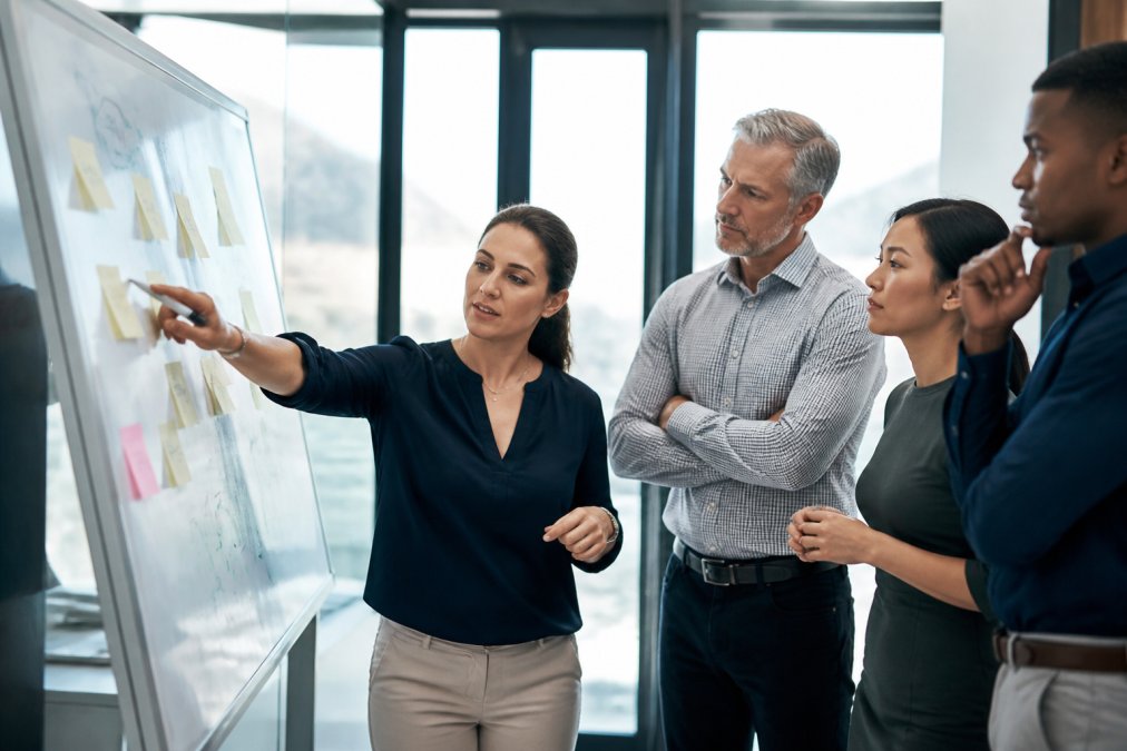 Team of managers gathered around a whiteboard in a modern office, collaborating on implementation plans with sticky notes and diagrams, natural daylight from large windows