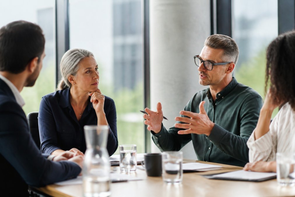 A diverse group of managers in a modern conference room engaged in active discussion, with one person listening intently while another gestures while speaking, natural lighting from large windows