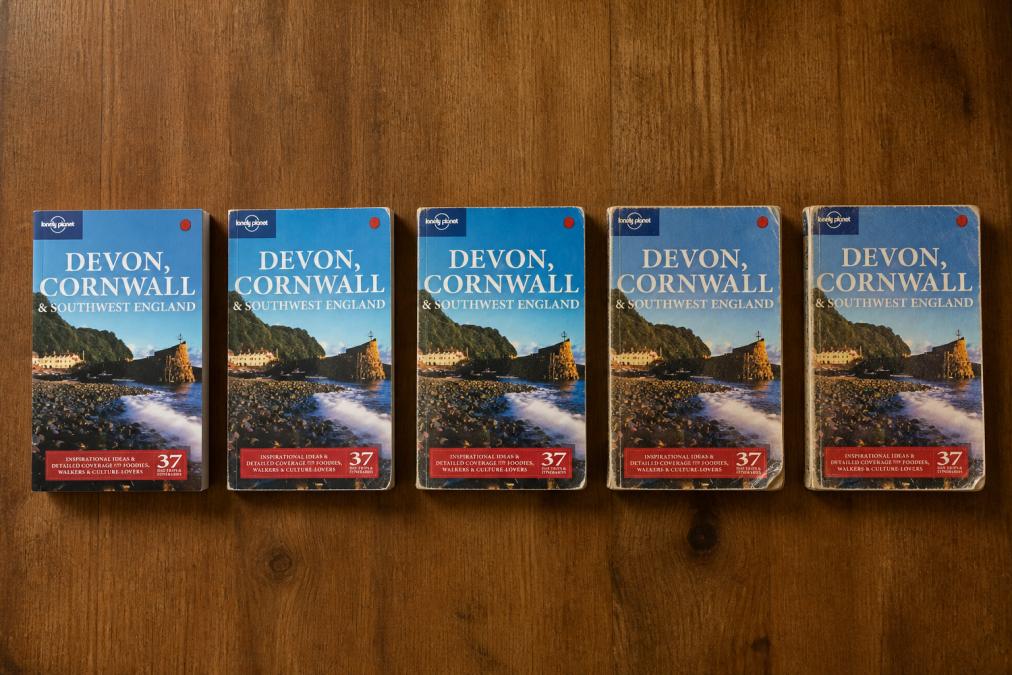 Overhead flat-lay of five second-hand books arranged showing progressively visible wear patterns, spine creasing, page yellowing, corner bending, demonstrating quality condition variations