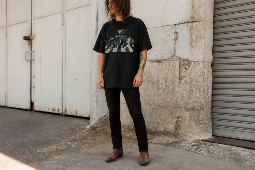 Person wearing an oversized vintage-inspired rock band graphic tee in black, styled with fitted dark jeans and vintage leather boots, standing in natural daylight showing how the oversized silhouette drapes naturally