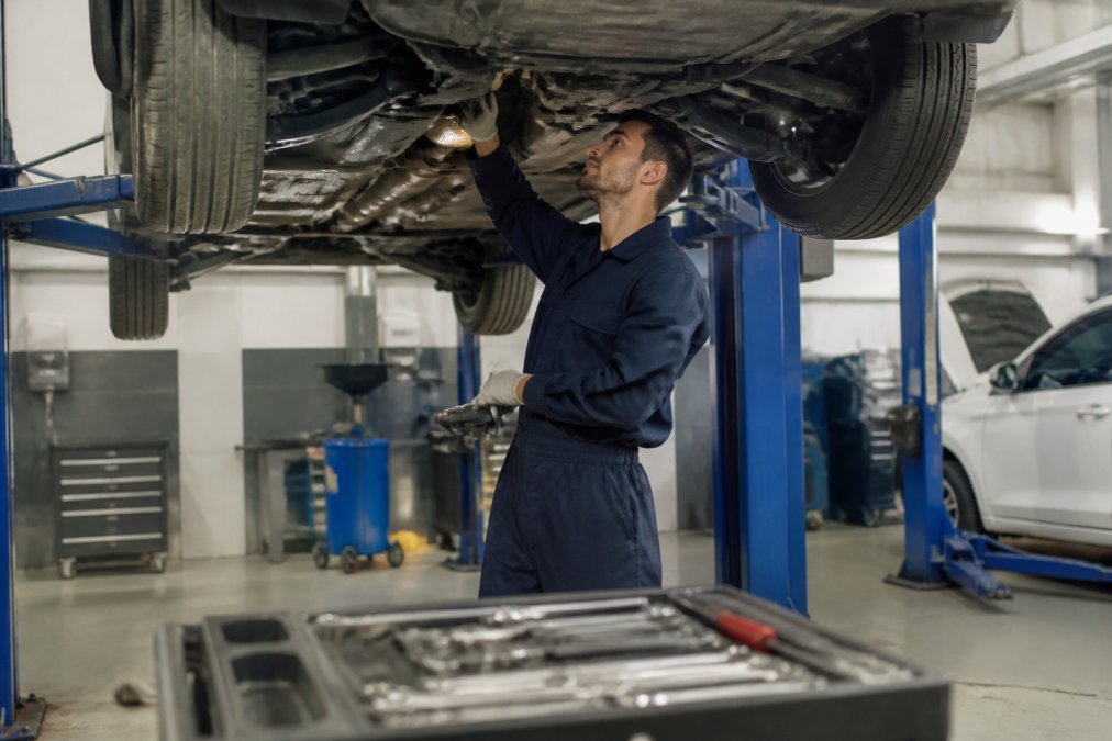 A certified mechanic in a clean, modern auto repair shop inspecting a vehicle raised on a lift, diagnostic tools visible in the foreground under bright overhead lighting