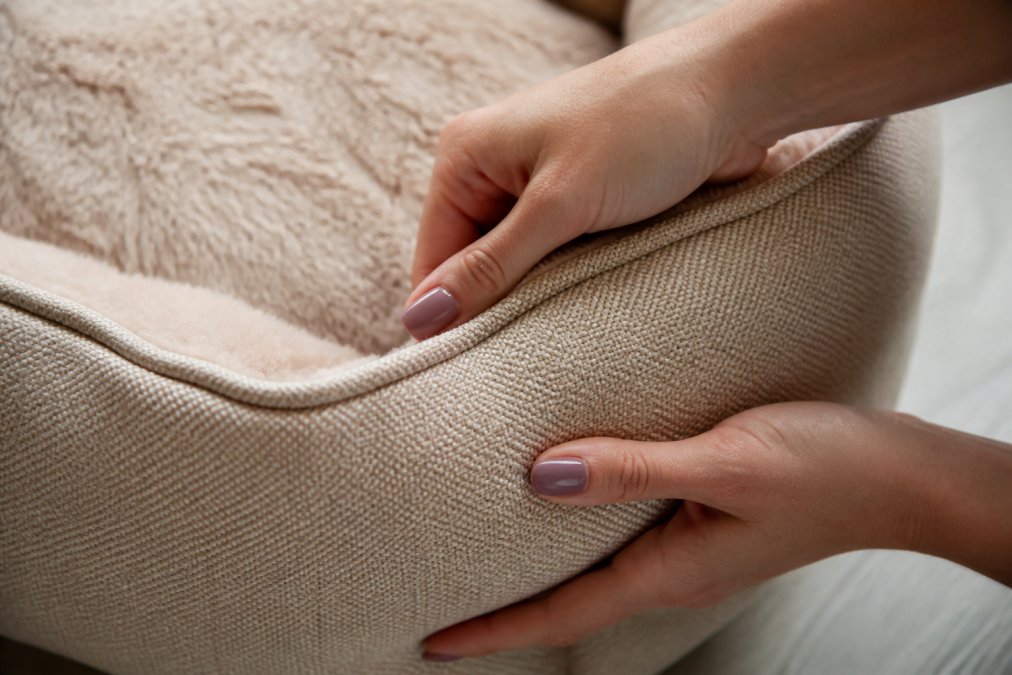 Close-up of hands examining a soft beige pet bed fabric, showing detailed stitching and texture quality under natural light