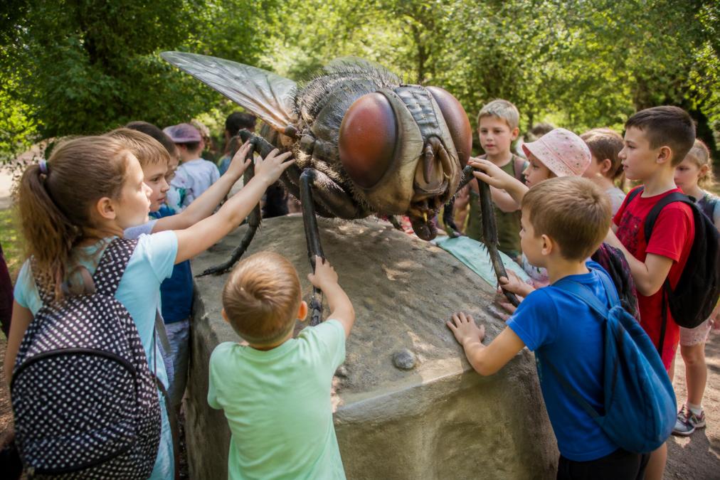 Grupa dzieci w wieku szkolnym stoi wokół gigantycznego modelu powiększonego owada na ścieżce edukacyjnej, dotykając i obserwując szczegóły figury, naturalne oświetlenie parku