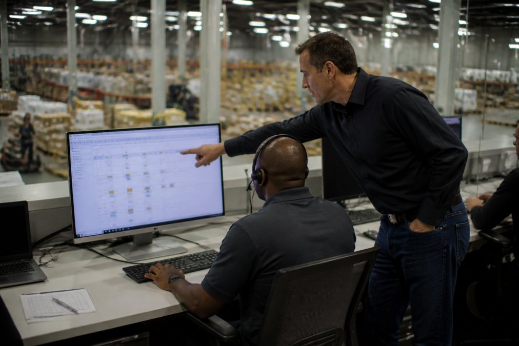 A logistics manager in a warehouse control room reviewing a dashboard on a large monitor, wearing a headset while a colleague points at a delivery schedule on the screen, fluorescent lighting over a busy operations floor