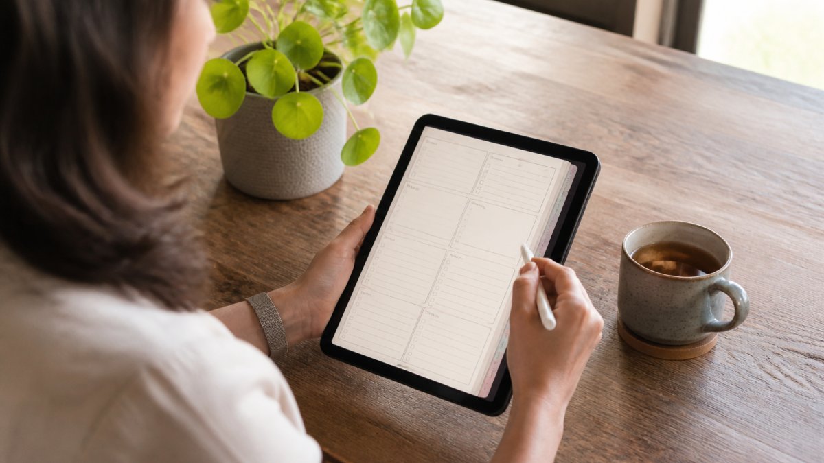 Person using a tablet with a stylus for digital planning at a sunlit desk with tea and a plant
