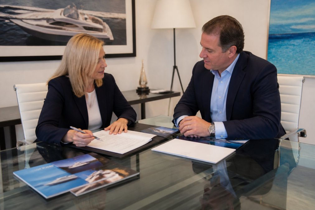 Two professionals in business attire engaged in focused discussion across a modern glass-topped desk in a bright office, with a framed yacht photograph and maritime artwork on the wall behind them