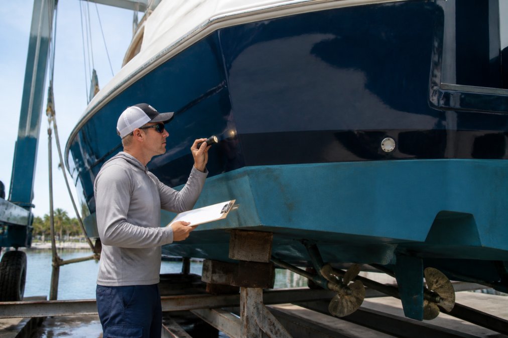 A marine surveyor in casual work clothes inspecting the hull of a large sportfish yacht on a lift, holding a clipboard and flashlight, with the yacht's lower hull visible and water in the background