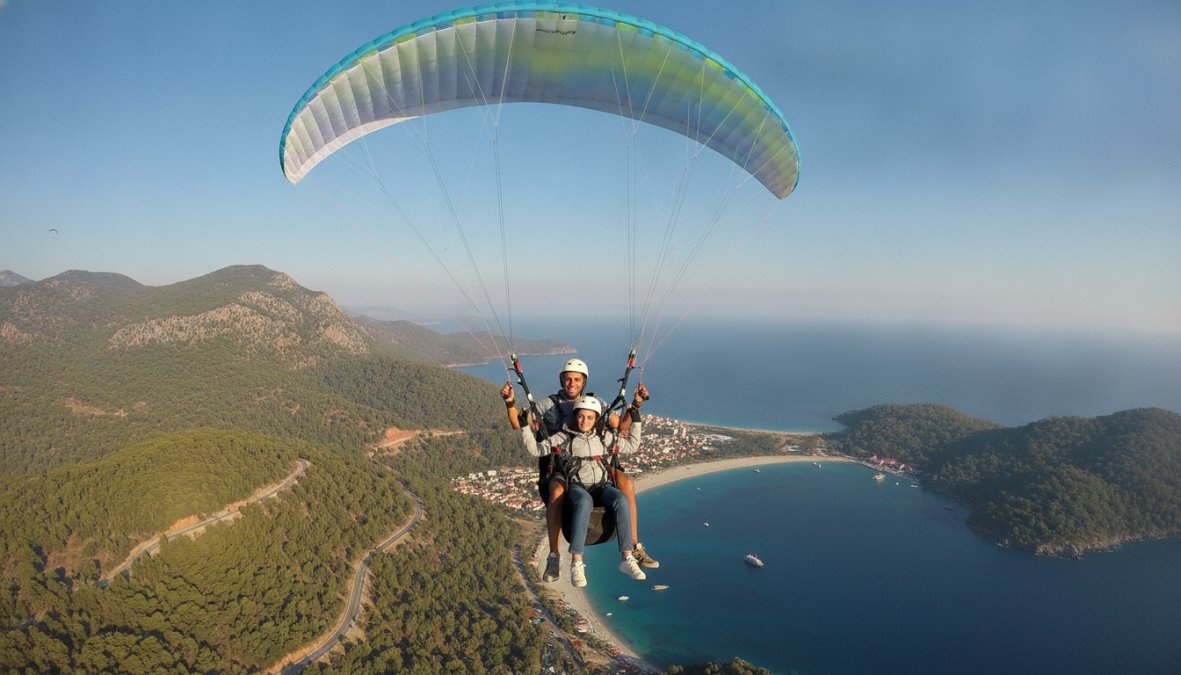 Deneyimli bir pilot ve gülümseyen yolcunun, renkli açık paraşütün altında gökyüzünde uçuş yaptığı, arka planda Fethiye'nin dağlık ve deniz manzarası görülen an