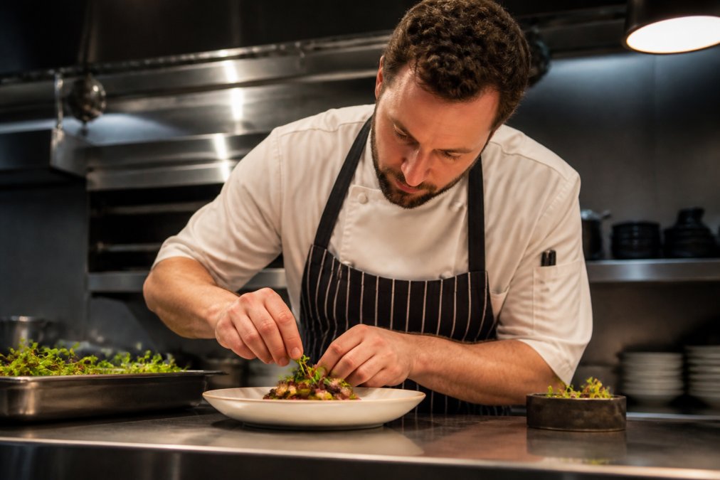Professional chef plating an elegantly prepared dish with precision, hands steady over a white ceramic plate in a modern kitchen with soft overhead lighting
