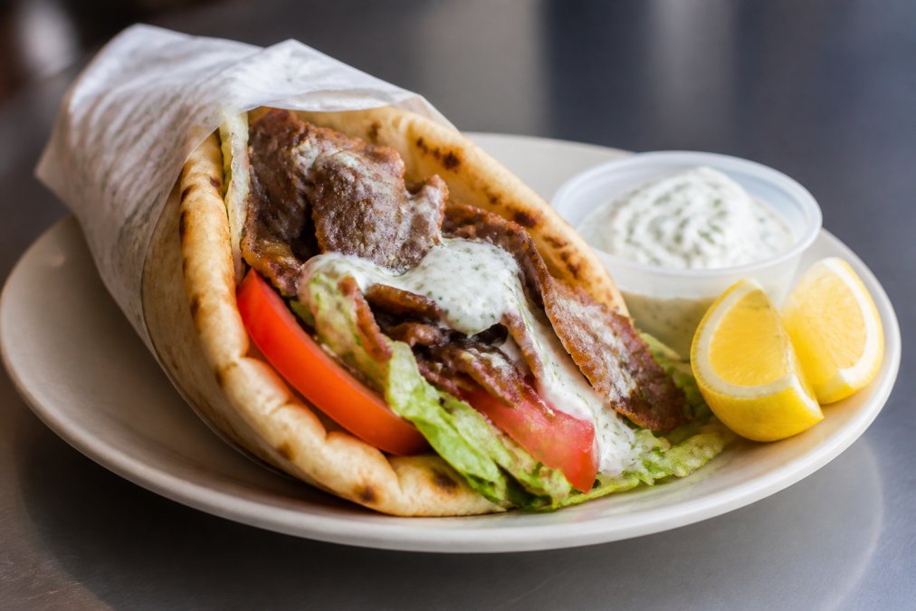 A freshly prepared gyro sandwich with seasoned meat, crisp lettuce, ripe tomato slices, and creamy white sauce wrapped in warm pita bread, served on a white plate with bright yellow lemon wedges and a small side of tzatziki sauce on the side
