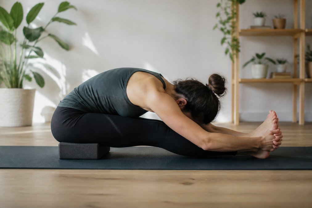 Person in seated forward fold position using a yoga block under their hips for support, demonstrating proper spinal alignment and hamstring engagement