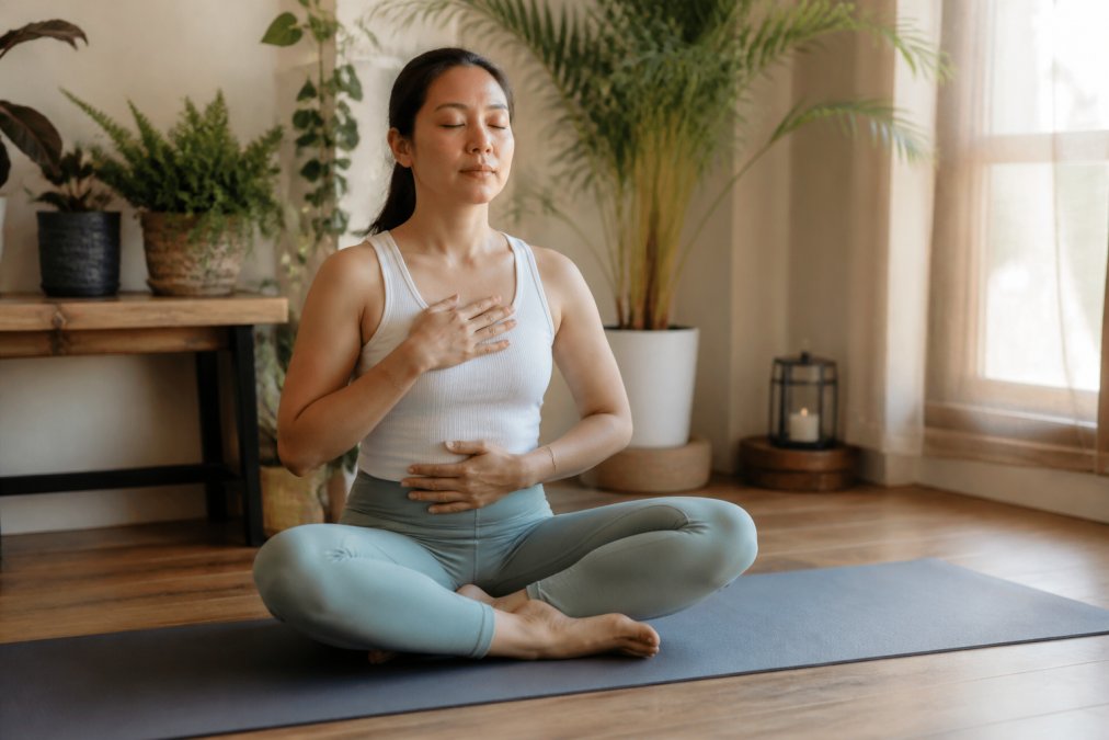 Person sitting cross-legged on yoga mat in peaceful home environment, eyes closed, hand on chest, practicing conscious breathing with calm expression and natural window lighting