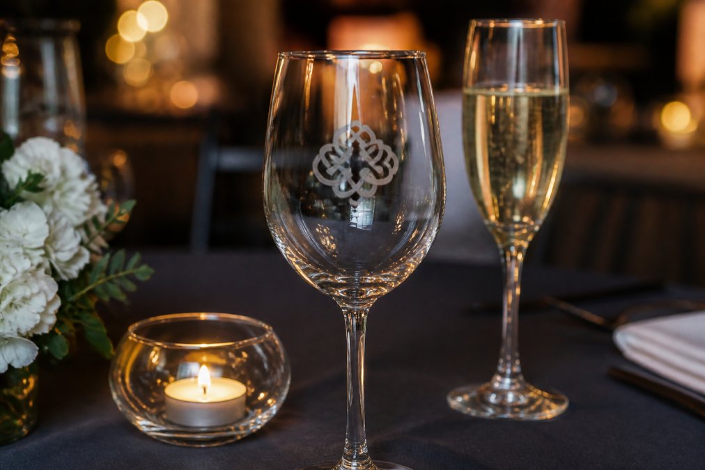 Elegantly etched wine glass with a corporate logo positioned on a hospitality venue table with soft candlelight highlighting the engraved detail, champagne flute beside it