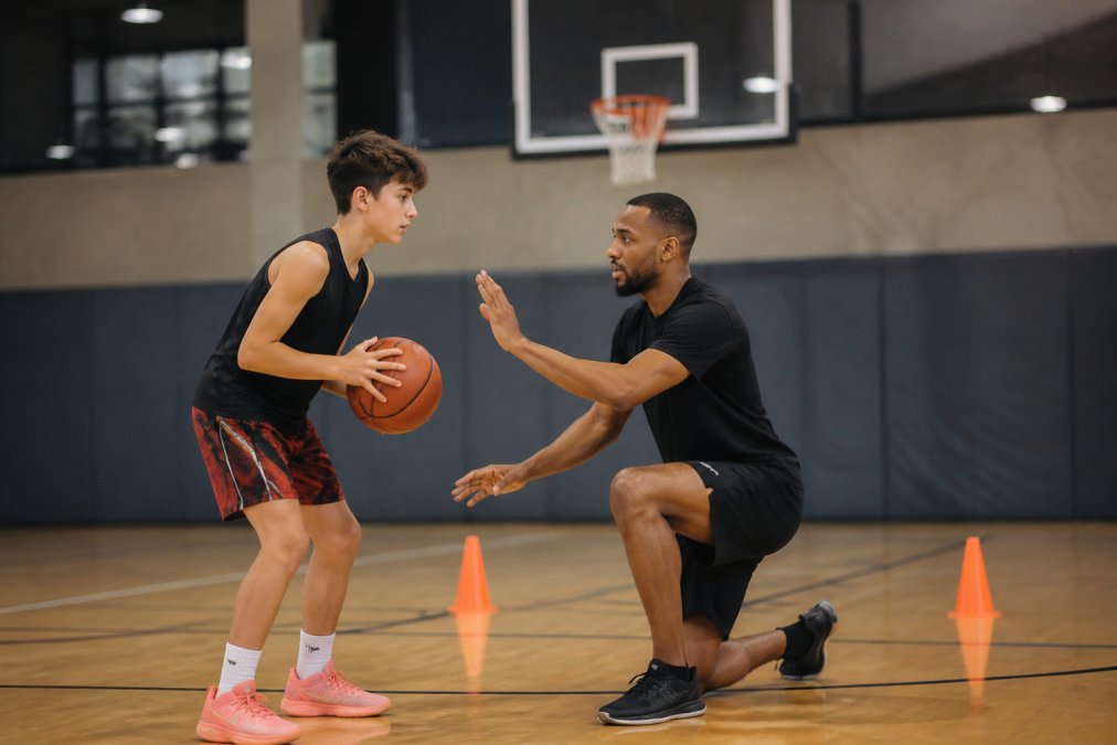 A coach working one-on-one with a young athlete on shooting mechanics and footwork during a training session, demonstrating proper form with focused attention in an indoor gymnasium