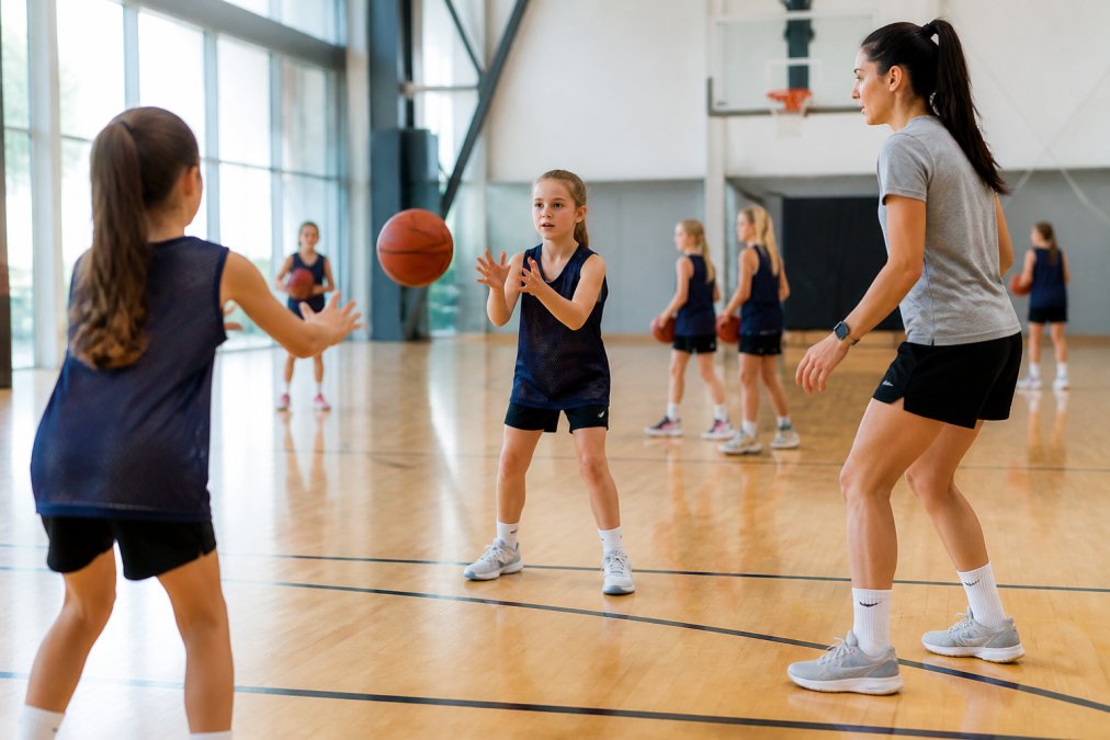 Young girls in basketball practice wearing team pinnies, executing passing drills with a coach demonstrating proper footwork on a wooden indoor court with natural gymnasium lighting