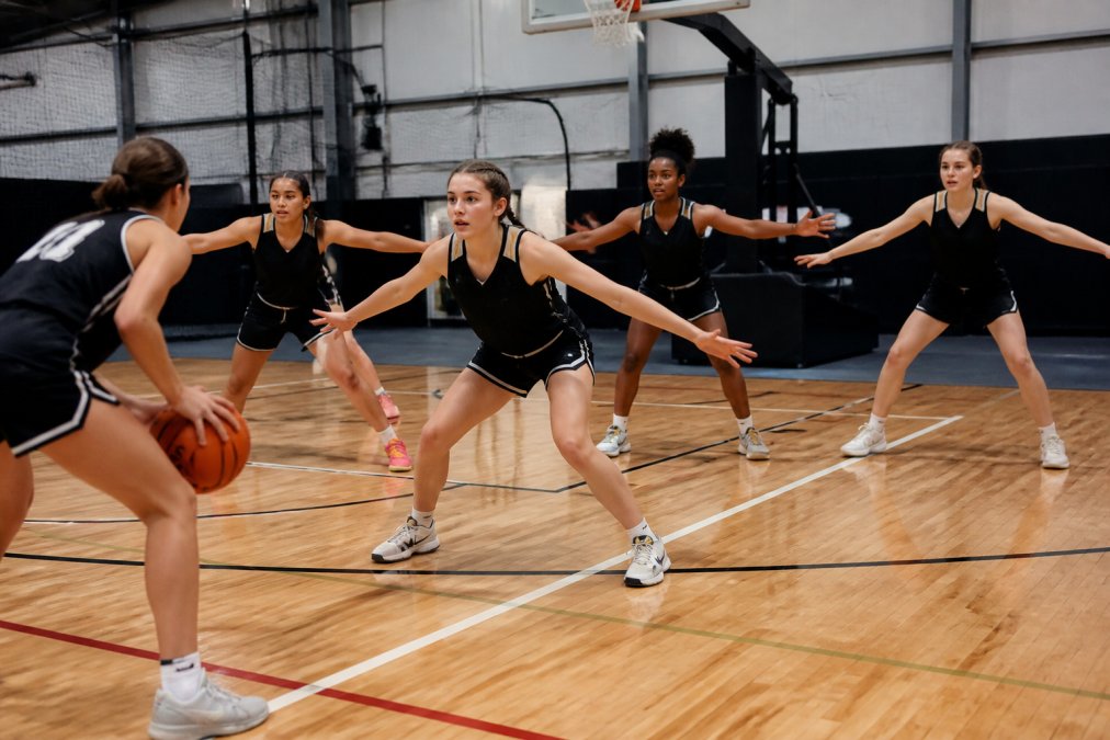 Diverse group of youth female basketball players in matching team uniforms executing a full-court press defensive drill with intensity and focus on a professional indoor court with bright overhead lighting