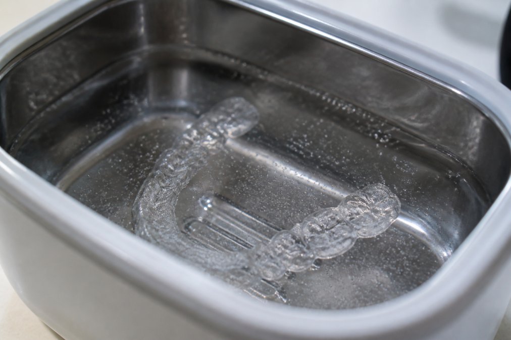 Nahaufnahme einer transparenten Zahnschiene, die in ein Ultraschallbad mit klarem Wasser gelegt wird, während feine Blasen um die Schiene herum sichtbar sind