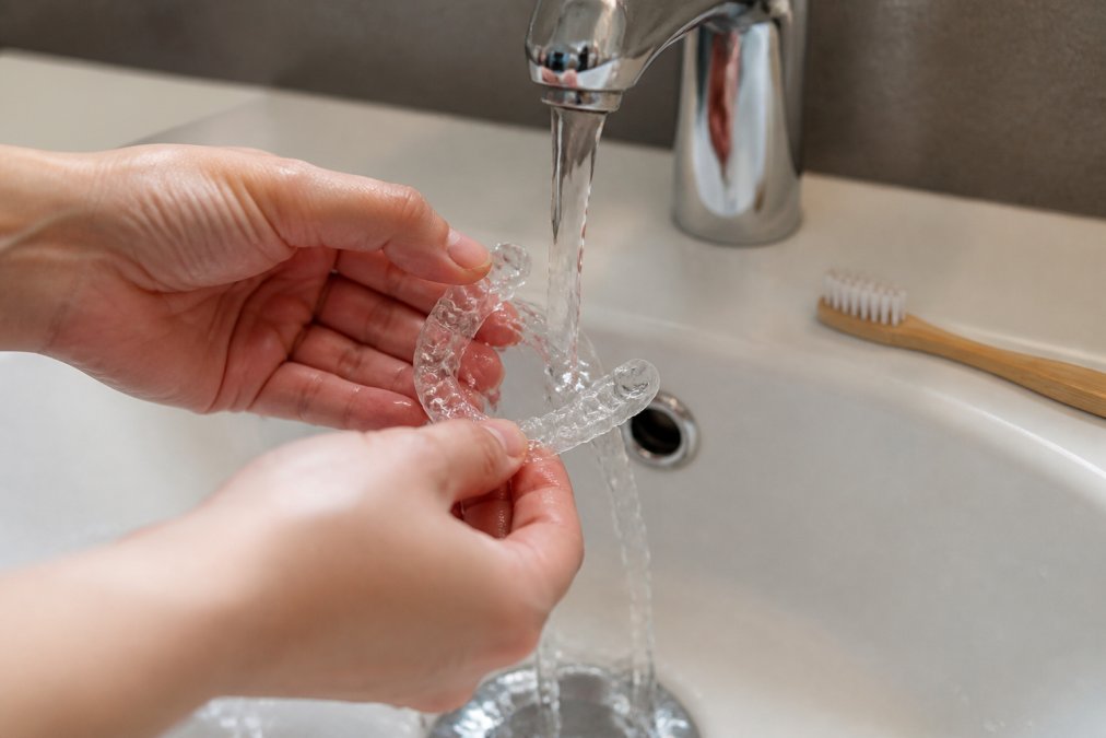 Nahaufnahme einer Zahnschiene unter fließendem lauwarmen Wasser, Person hält die Schiene vorsichtig in den Händen über einem Waschbecken, eine weiche Zahnbürste liegt daneben auf dem Beckenrand