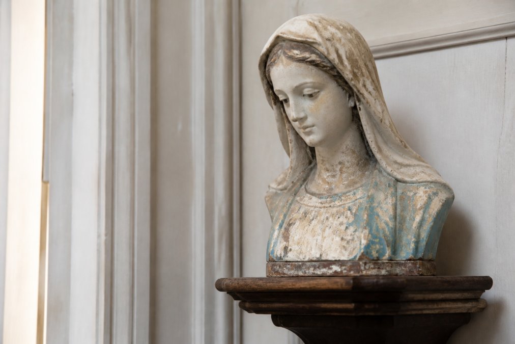 Close-up van een antiek gipsen Mariabeeld met serene gelaatsuitdrukking op een houten console, zachte natuurlijke belichting die de patina en verfdetails benadrukt