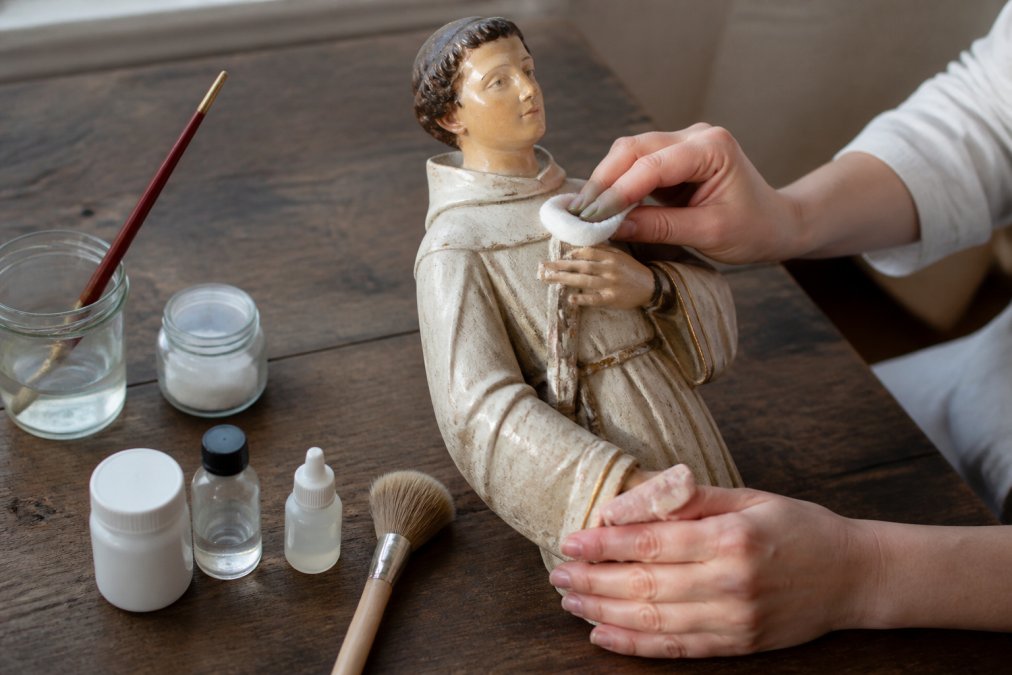 Handen die voorzichtig een antiek gipsen heiligenbeeld reinigen met een zachte doek op een houten tafel, omringd door conserveringsmiddelen en een kwastje