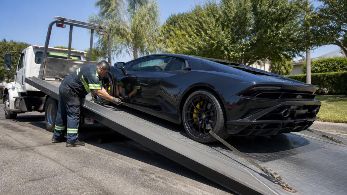 Professional towing operator positioning a low-clearance exotic car onto a flatbed truck with hydraulic ramp at safe shallow angle, showing soft straps securing the vehicle and tie-down placement in detail