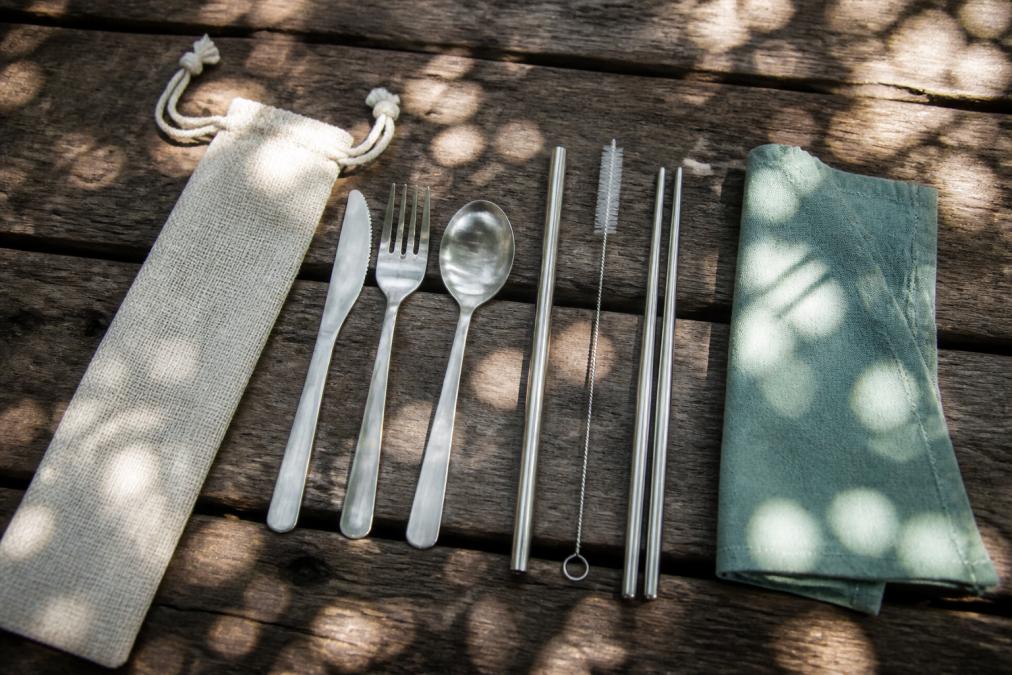 Overhead shot of a stainless steel travel cutlery set laid out on a weathered wooden picnic table beside a hemp pouch, reusable straw, chopsticks, and a cloth napkin, dappled afternoon sunlight through tree leaves