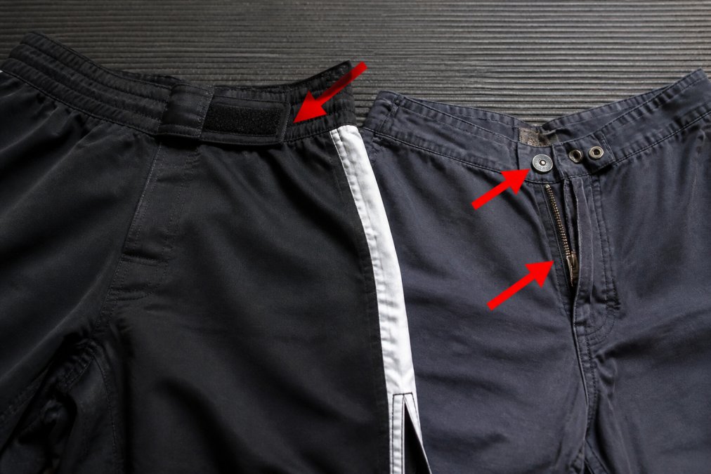 Close-up van MMA shorts met klittenbandsluiting en gladde naden, naast normale sportbroek met rits en knopen, met pijlen die de scherpe onderdelen aanwijzen