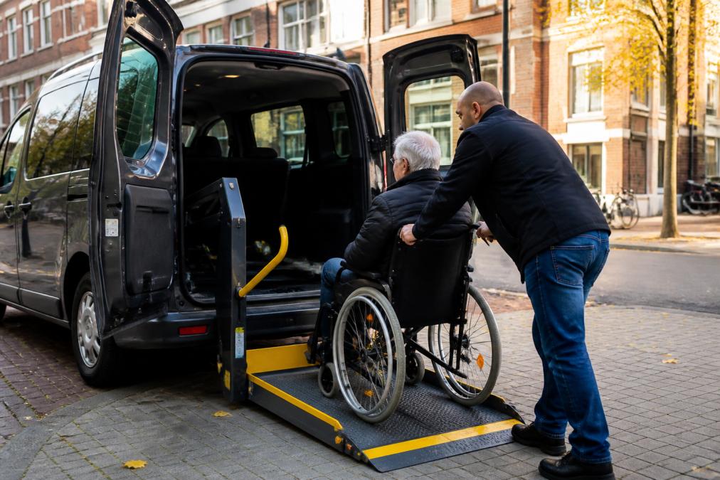 Gespecialiseerde rolstoeltaxi met open deuren, zichtbaar aangepaste lift en gele veiligheidsbeugels, professionele chauffeur helpt oudere rolstoelgebruiker voorzichtig in de auto op een zonnige straathoek in Utrecht