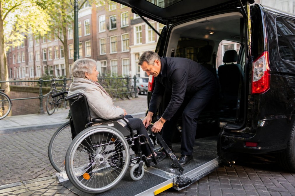 Een vriendelijke chauffeur helpt een oudere vrouw in een rolstoel veilig de rolstoeltaxi in te rijden via een oprijplaat, terwijl hij de rolstoel fixeert voor vertrek. De chauffeur draagt een net uniform en de omgeving is een rustige straat in Utrecht met zacht ochtendlicht.
