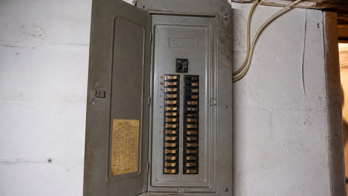 Close-up of a Zinsco electrical panel mounted on a basement wall, showing distinctive tan-colored breaker switches, the Zinsco branding on the panel cover, and the characteristic rectangular shape of vintage 1970s-era panels