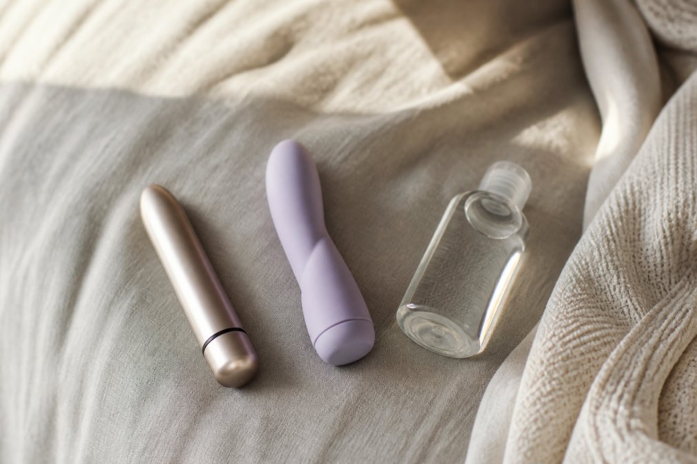 Een elegante, minimalistische bullet vibrator en een compacte vibrator liggen op een zachte, neutraal gekleurde stoffen ondergrond naast een kleine fles glijmiddel op waterbasis. De setting is rustig en sfeervol met zacht natuurlijk licht.
